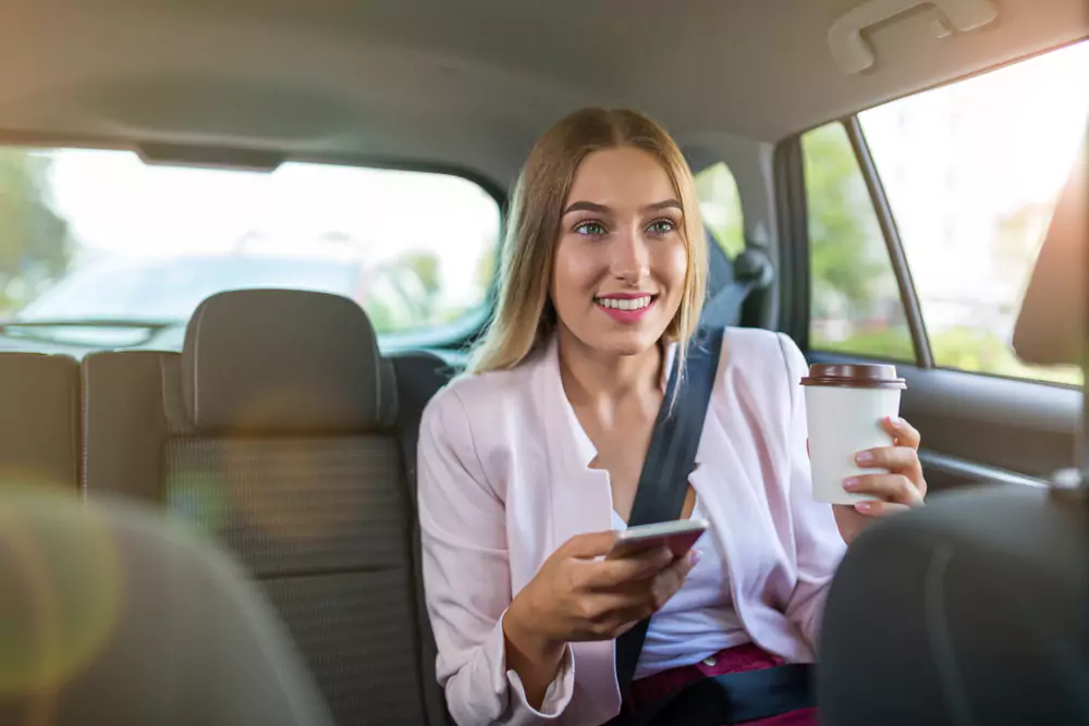 "Smiling woman seated in the back of a car, holding her phone in her right hand and a cup of coffee in her left hand. She appears joyful, suggesting she's enjoying a taxi ride."