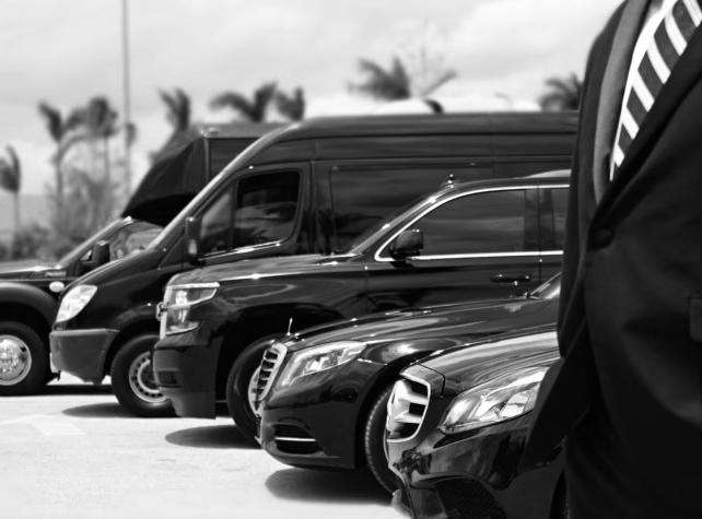 A grayscale image featuring a lineup of vehicles, ranging from a small car to a bus, with a man in a black suit standing in front. The man's body is obscured, and only the lower part of the vehicles is visible.